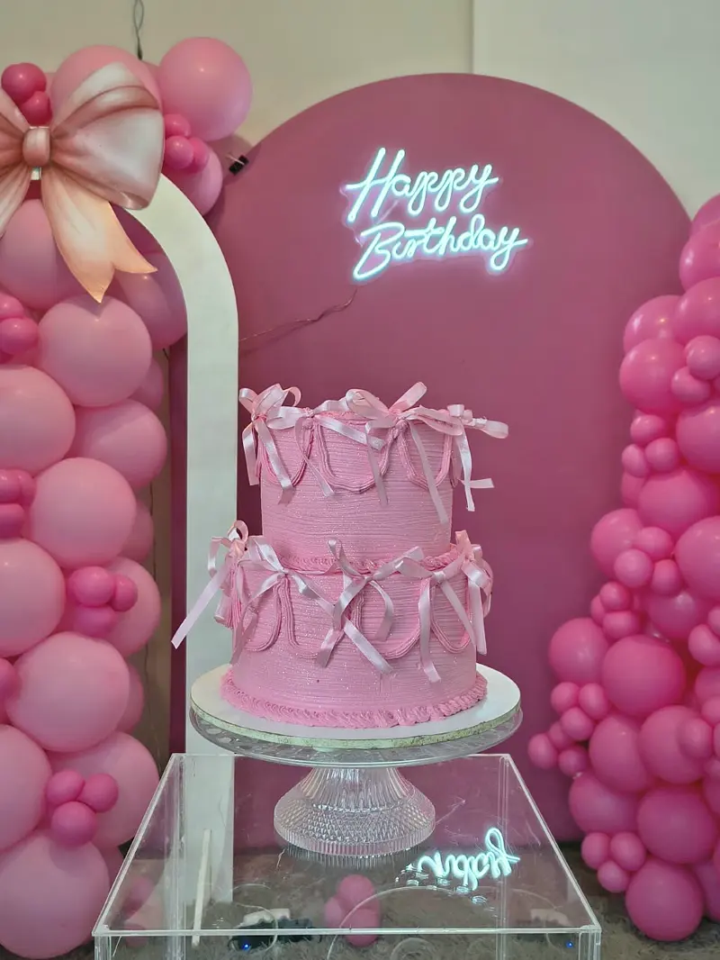 Bolo rosa de dois andares com laços em frente ao letreiro Happy Birthday